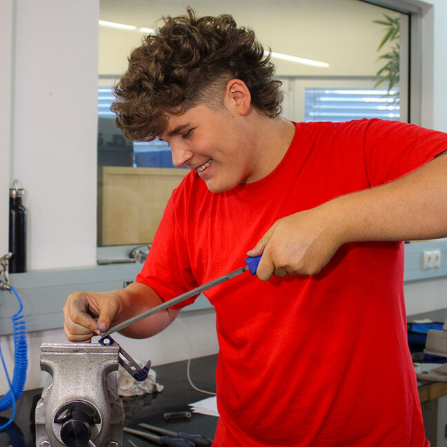 Ein Metalltechnik-Lehrling führt präzise Feilarbeiten an einem eingespannten Metallwerkstück durch. Die Aufnahme zeigt die praktische Ausbildung in der Metallbearbeitung mit Handwerkzeugen in einer modernen Werkstatt. Zu den typischen Tätigkeiten zählen Feilen, Anpassen und Bearbeiten von Metallbauteilen für Maschinen- und Anlagenkomponenten.